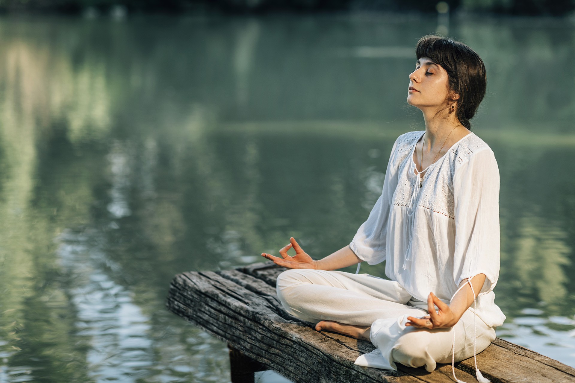 Yoga Retreat. Peaceful young woman sitting in lotus position and meditating by the lake